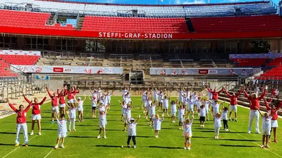 In front of a red grandstand, children and adults in red and white sportswear stand in rows and raise both arms into the air.